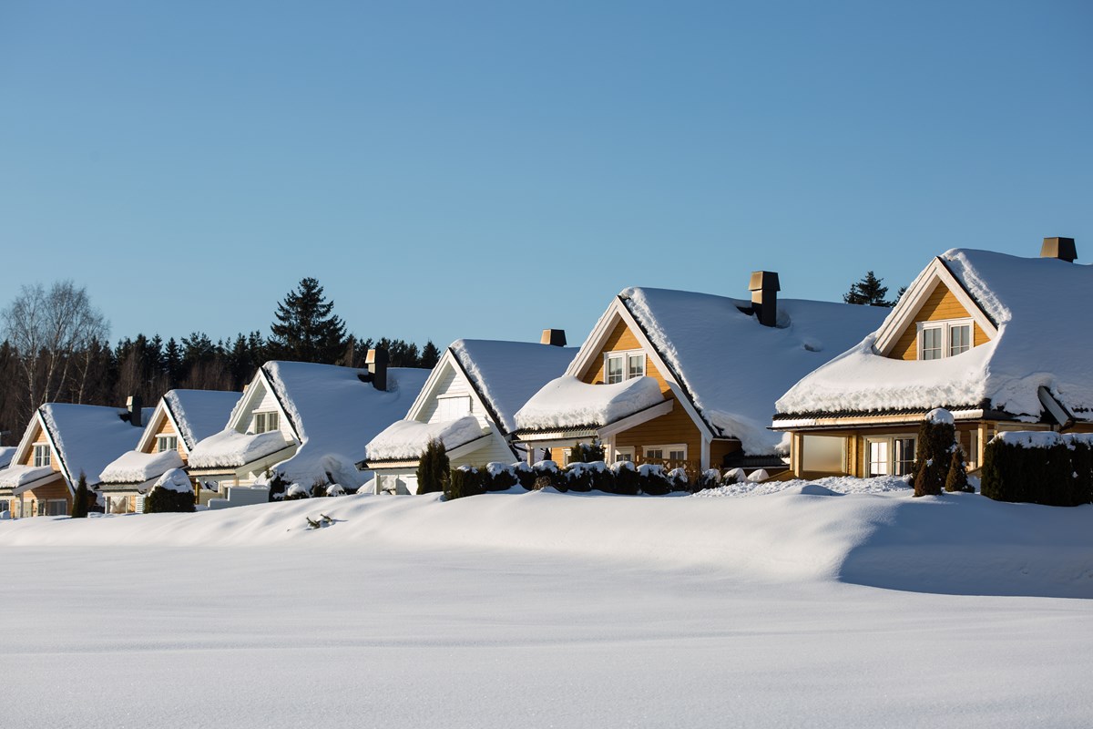 Hus dekket av dyp snø, rommet av blå himmel og sol som skinner mot husveggen. Foto.