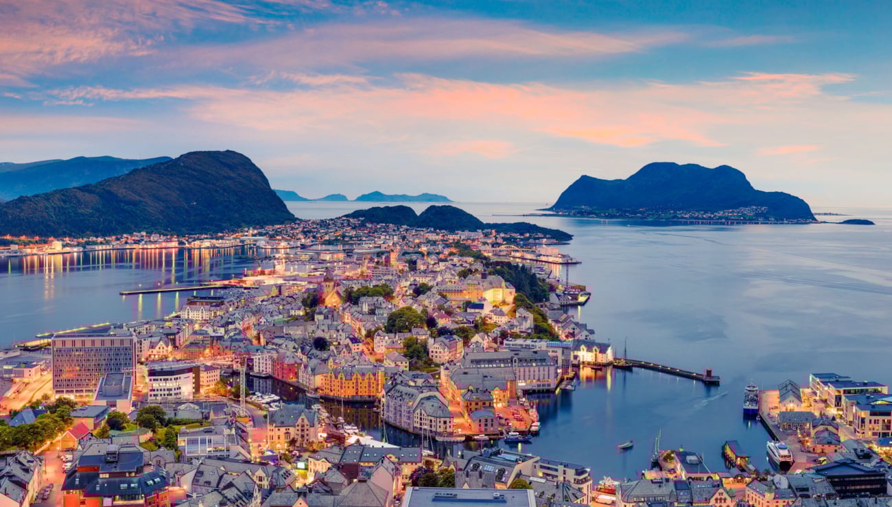 Utsikt over Ålesund by og fjorden i skumring. Himmelen er rosa, og det lyser over byen. Foto.