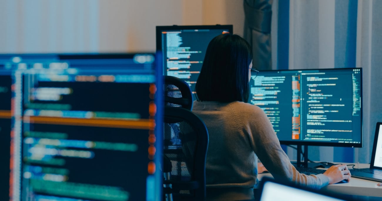 Et kontorlandskap med store pc-skjermer og en kvinne som arbeider med koding. Foto.